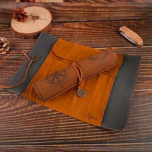 May include: A brown leather pen roll with a compass design and a world map on the outside. The roll has a black canvas flap with the word "Aspen" embossed on it.