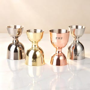 May include: Four stainless steel jiggers in silver, gold, and copper tones. Each jigger has a flared top and a bulbous base. The gold jigger has "RP" engraved, the copper one has "P.W.F", and the silver one has "JM".