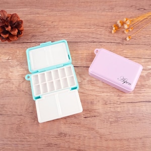 以下が含まれることがあります： 空のウォーターカラーペイントパレットが2つ。1つは薄い青で、15個のくぼみとヒンジ付きの蓋が付いています。もう1つは薄い紫で、ヒンジ付きの蓋が付いており、前面に「AiPen」と印刷されています。