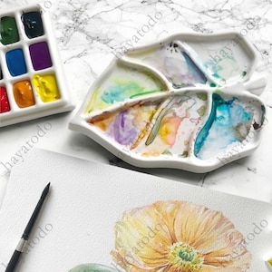 以下が含まれることがあります： カラフルな絵の具の残骸が付いた、白いセラミック製の葉の形をした水彩パレット。さまざまな色の絵の具が入った白い長方形のパレットが見えます。黄色の花の水彩画と筆も画像にあります。