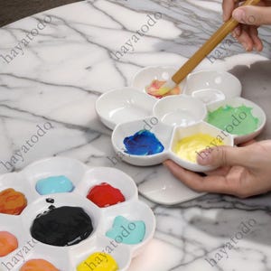 以下が含まれることがあります： 絵の具を混ぜるための複数のくぼみがある、白いセラミック製の絵の具パレットが2つ。パレットには、青、緑、黄、赤、オレンジ、黒など、さまざまな色の絵の具が入っています。