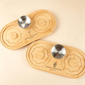 以下が含まれることがあります： ステンレス製のボウルが付いた2つの竹製サービングトレイ。各トレイには花の模様と「MARIA JOHNSON bartender」の文字が入っています。楕円形のトレイは天然木仕上げです。