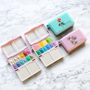 以下が含まれることがあります： ピンクとティール色のケースに入った、小さな水彩絵の具セットのコレクション。各ケースには花の模様とテキストが施されています。開いたセットからは、さまざまな鮮やかな色が見えます。ケースは大理石の表面に置かれています。
