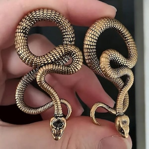 May include: A pair of gold-toned snake-shaped ear gauges. The gauges are made of metal and have a detailed, textured surface. The snakes are curled into a circular shape and have black eyes.