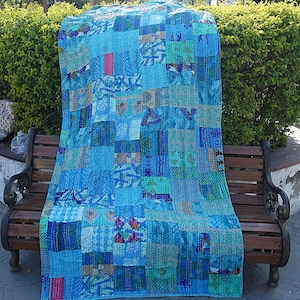 May include: A patchwork quilt made from repurposed fabric in shades of blue, green, and pink. The quilt is made up of many different squares and rectangles, each with a unique pattern. The quilt is folded and displayed on a wooden bench.