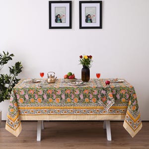 May include: A rectangular tablecloth with a floral pattern in shades of green, pink, and yellow. The tablecloth is set on a white table with a teapot, plates, and a vase of flowers. Two framed pictures hang on the wall.