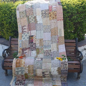 Op de afbeelding: Een patchworkdeken gemaakt van verschillende stoffen in tinten bruin, beige en wit. De deken is gevouwen en over een houten bank gelegd.