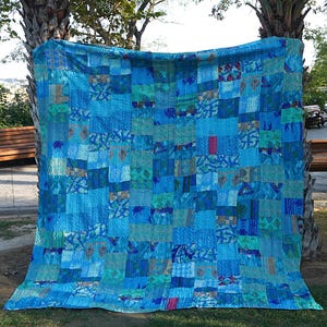 May include: A large blue patchwork quilt made from many different fabrics. The quilt is made up of squares and rectangles of various sizes and patterns. The quilt is hanging on a clothesline in a garden setting.