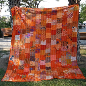 May include: A large, orange and red patchwork quilt made from recycled saris. The quilt is made up of many different squares of fabric, each with a unique pattern. The quilt is perfect for adding a touch of color and warmth to any room.
