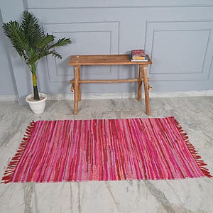 Op de afbeelding: Een gestreept geweven tapijt in roze en rood met franjes aan de randen. Het tapijt ligt op een lichtbruine tegelvloer voor een houten bank.