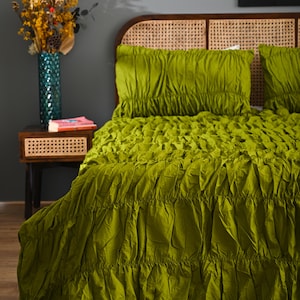 Op de afbeelding: Een bed met een groene, gerimpelde dekbedovertrek en bijpassende kussenslopen. Het hoofdbord is gemaakt van geweven rotan. Een kleine houten nachtkastje met een vaas met bloemen en een boek is zichtbaar. Thema: interieurdecoratie.