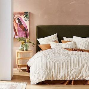 Op de afbeelding: Een slaapkamer met een groen hoofdbord en wit beddengoed met een boogpatroon. Decoratieve kussens in wit en bruin passen bij het bed. Een houten nachtkastje met een geweven voorkant en een schilderij dragen bij aan de inrichting van de kamer.