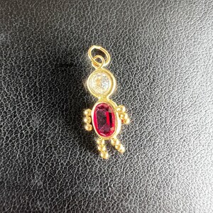 May include: A gold-colored pendant in the shape of a person. The head is a circle with a clear stone, the body is an oval-shaped red stone, and the arms and legs are small gold balls. The pendant is on a black surface.