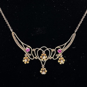 May include: A gold-toned necklace with a delicate filigree design. The necklace features heart-shaped elements, pink gemstones, and small flower-shaped accents. The necklace is displayed against a black background.