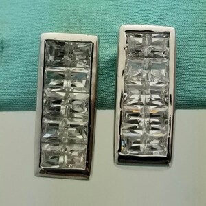 May include: A pair of rectangular silver-toned earrings. Each earring features a series of clear, square-cut stones set within a rectangular frame. The stones are arranged in a grid pattern, creating a sparkling effect. The earrings are displayed against a light blue background.