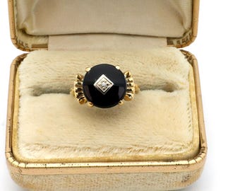 Könnte beinhalten: Ein goldener Ring mit einem großen, runden schwarzen Onyxstein und einem kleinen, quadratischen Diamanten in der Mitte. Der Ring ist in einem goldenen Band mit dekorativen Akzenten gefasst. Präsentiert in einer Vintage-Schmuckschatulle in Gold mit cremefarbenem Samt.