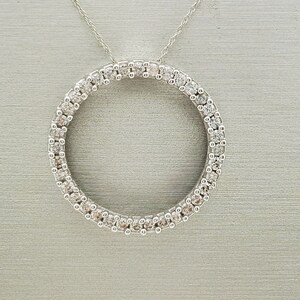 May include: A silver necklace featuring a circular pendant encrusted with numerous small, clear gemstones. The pendant is suspended from a delicate silver chain, creating a minimalist and elegant design. The gemstones sparkle, adding a touch of glamour.