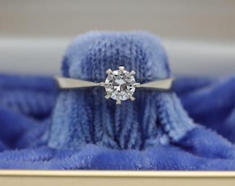 May include: A solitaire diamond ring with a round-cut diamond set in a silver band. The ring is displayed on a blue velvet surface, inside a jewelry box. The diamond is the focal point, sparkling brightly.