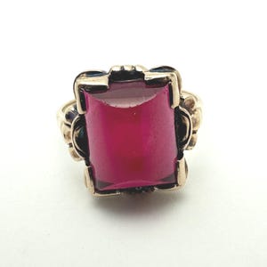 May include: A gold ring featuring a large, rectangular, deep pink gemstone set in a prong setting. The metal band has decorative, scalloped edges surrounding the central stone, creating a bold and structured appearance for this piece of jewelry.
