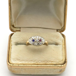 May include: A vintage gold ring with an oval cluster of diamonds, a blue gemstone, and a red gemstone, set in a gold band. The ring is displayed in a vintage, gold-toned jewelry box with a cream-colored velvet interior.