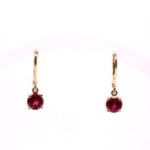 May include: A pair of drop earrings featuring round, faceted, deep pink stones set in gold-toned metal. Each earring has a hinged lever-back closure that connects to a small circular frame holding the vibrant, translucent gemstone securely in place.