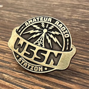 May include: A brass-colored pin with the words "AMATEUR RADIO" arched across the top. Below, the text "W5SN STATION" is displayed on a banner. The pin features a radio tower and lightning bolt design.