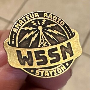 May include: A gold-colored pin with the text "AMATEUR RADIO" arched at the top and "W5SN STATION" at the bottom. The center features a radio tower with lightning bolts. The pin is a vintage style.