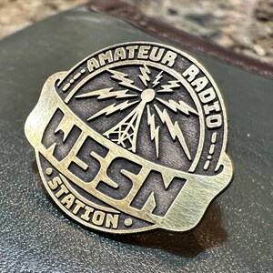 May include: A bronze-colored pin with the words "AMATEUR RADIO" and "WSSN STATION" encircling a central design of a radio tower and lightning bolts. The pin is set against a textured, dark green surface.