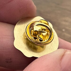 May include: A gold-colored lapel pin with a round, scalloped edge. The pin features a gold-colored clasp on the back for secure attachment. The pin is held between two fingers.