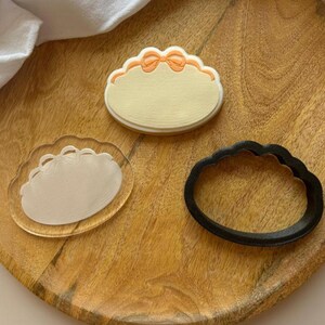 Puede incluir: Un conjunto de tres artículos para decorar galletas: una galleta beige con un diseño de lazo naranja, un sello acrílico transparente con el contorno de un lazo y un cortador de galletas negro con forma de óvalo festoneado con un lazo.