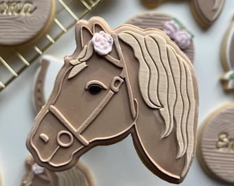 May include: A light brown horse-shaped sugar cookie with a dark brown eye, a tan bridle, and a small pale pink fondant rose on the mane. The cookie features embossed details on the mane and face, presented among other decorated cookies on a wire rack.