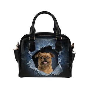 Puede incluir: Bolso de mano negro con un diseño de mezclilla desgastada que presenta un perro marrón asomándose a través de una sección rasgada. El bolso tiene asas negras, una correa para el hombro y herrajes dorados.