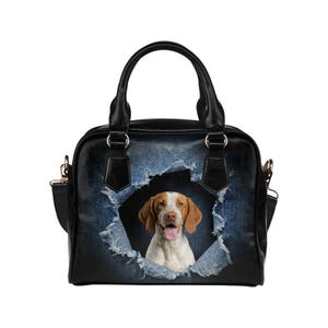 May include: Black handbag with a distressed denim design featuring a dog peeking through a torn hole. The dog has brown and white fur and is smiling. The bag has a top handle and a detachable shoulder strap.
