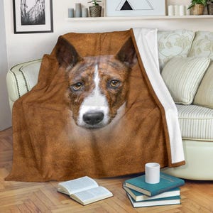 May include: A brown and white dog's face is featured on a throw blanket. The blanket is draped over a sofa, with the dog's face centered. The dog has brown eyes, a white snout, and brown and white fur. The blanket has a brown background.