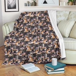 May include: A soft, plush blanket featuring a repeating pattern of smiling Staffordshire Bull Terrier faces in various shades of brown, black, and tan. The blanket is draped over a sofa, with a white backing visible.