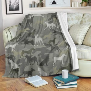 May include: A gray and white dog-themed blanket with a camouflage pattern. The blanket features silhouettes of dogs in various poses, creating a playful and unique design. The blanket is draped over a sofa.