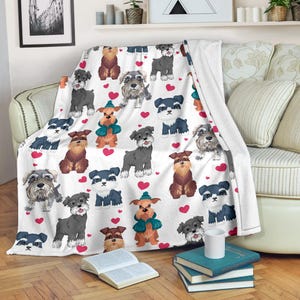 Op de afbeelding: Witte plaid met een herhaald patroon van Schnauzer hondenillustraties in verschillende kleuren, afgewisseld met rode harten. De deken is over een bank gedrapeerd en toont het ontwerp en de textuur.