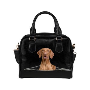 May include: Black handbag featuring a brown dog graphic peeking out from a zipper design. The bag has a structured shape with top handles and a detachable shoulder strap. Gold-colored hardware accents the bag's design.