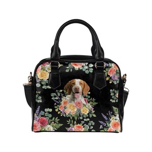 May include: Black handbag with a floral pattern and a dog portrait. The bag features a dog's face surrounded by colorful roses, daisies, and other flowers. It has black handles and a shoulder strap.