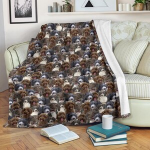 May include: A cozy blanket featuring a repeating pattern of smiling dog faces in various shades of brown, black, and white. The blanket is draped over a sofa, with a white edge visible. The blanket is made of soft material.