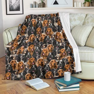 Può includere: Una morbida coperta con un motivo ripetuto di facce di cani Cocker Spaniel nei toni del nero, marrone e beige. La coperta è drappeggiata su un divano, con un retro bianco visibile. Libri e una tazza in primo piano.