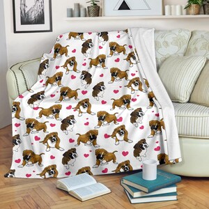 May include: White throw blanket featuring a repeating pattern of brown and white Boxer dogs and pink hearts. The blanket is draped over a sofa, showcasing the design and soft texture. A cozy home decor item.