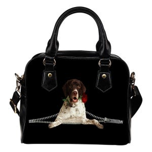 May include: Black handbag with a dog graphic. The dog is brown and white, holding a red rose in its mouth, and appears to be emerging from a zipper. The bag has a top handle and a detachable shoulder strap. The hardware is gold-colored.