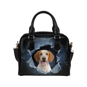 May include: Black handbag with a dog graphic. The bag features a distressed denim design with a torn effect, revealing a dog's face. It has a top handle and a detachable shoulder strap. The dog has brown and white fur.
