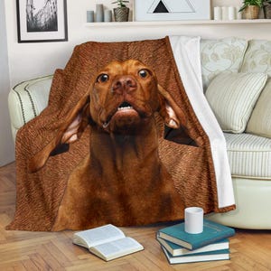 May include: A brown dog with large, floppy ears is featured on a throw blanket. The blanket is draped over a sofa, with a white border. The dog's surprised expression is captured in detail. The blanket is a warm, inviting home accessory.