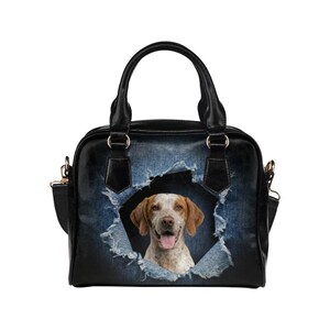 Könnte beinhalten: Schwarze Handtasche mit einem Design aus zerrissenem Denim, das einen Hund zeigt, der durch ein Loch blickt. Der Hund hat braunes und weißes Fell und einen fröhlichen Ausdruck. Die Tasche hat schwarze Griffe und einen Schultergurt.