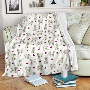 May include: White throw blanket featuring a repeating pattern of light-colored dogs and pink hearts. The blanket is draped over a sofa and is made of a soft, cozy material. A decorative item for the home.