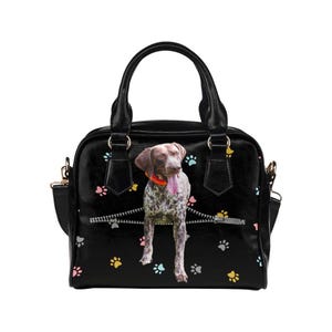 May include: Black handbag featuring a German Shorthaired Pointer dog graphic emerging from a zipper design. The bag has a top handle, a shoulder strap, and colorful paw prints scattered across the surface. The dog is wearing an orange collar.