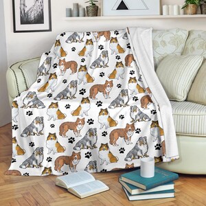 Puede incluir: Manta blanca con un patrón repetido de ilustraciones de perros Shetland y huellas de patas negras. Los perros están representados en tonos de marrón, gris y blanco. La manta está sobre un sofá.