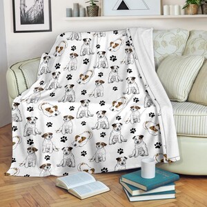 Peut inclure: Plaid blanc avec un motif répété d'illustrations de Jack Russell Terriers et d'empreintes de pattes noires. Les chiens sont représentés dans diverses poses, avec des marques marron et blanches. La couverture est drapée sur un canapé.